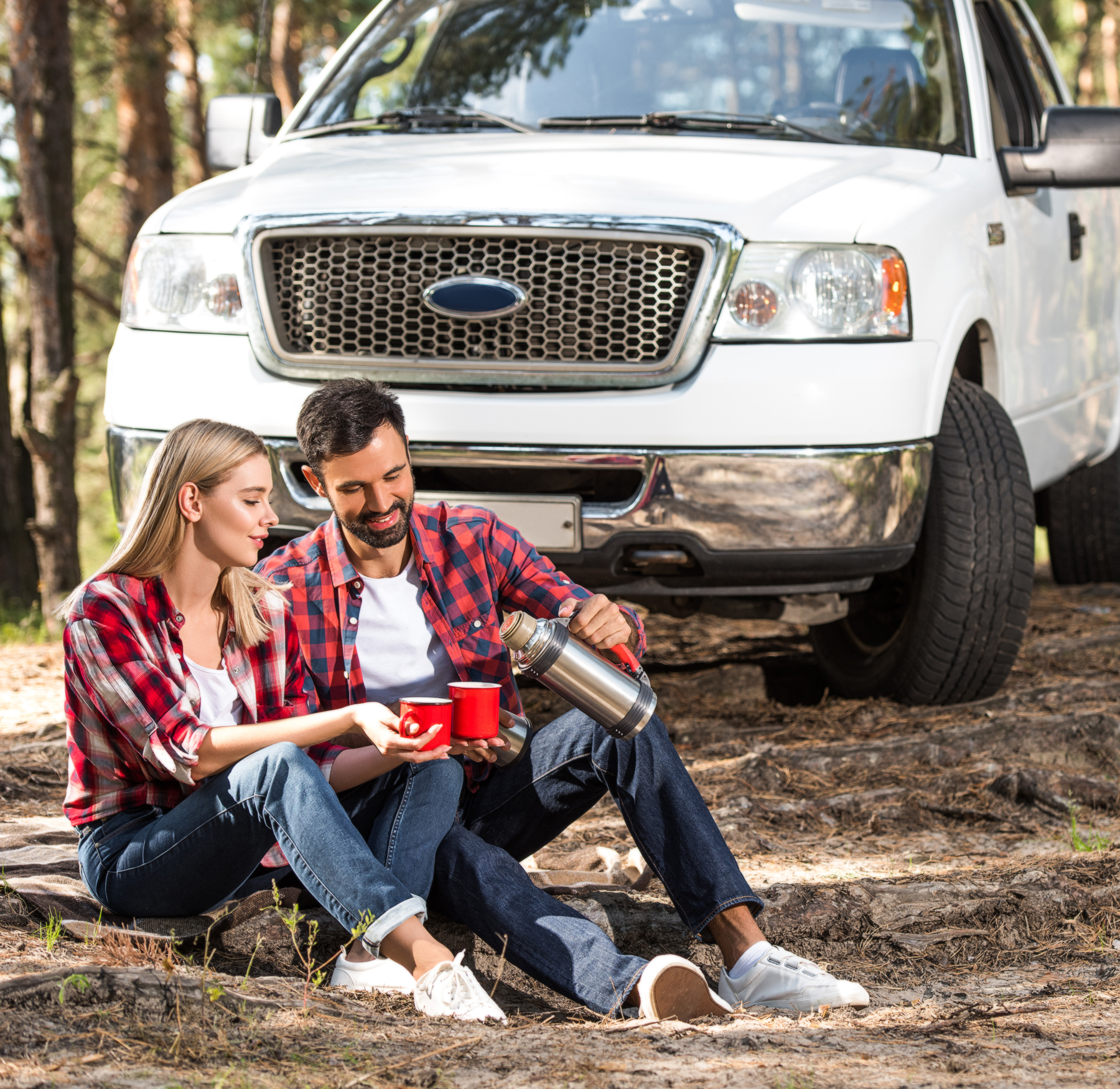 young couple sitting on ground drinking coffee in woods with white truck in the background