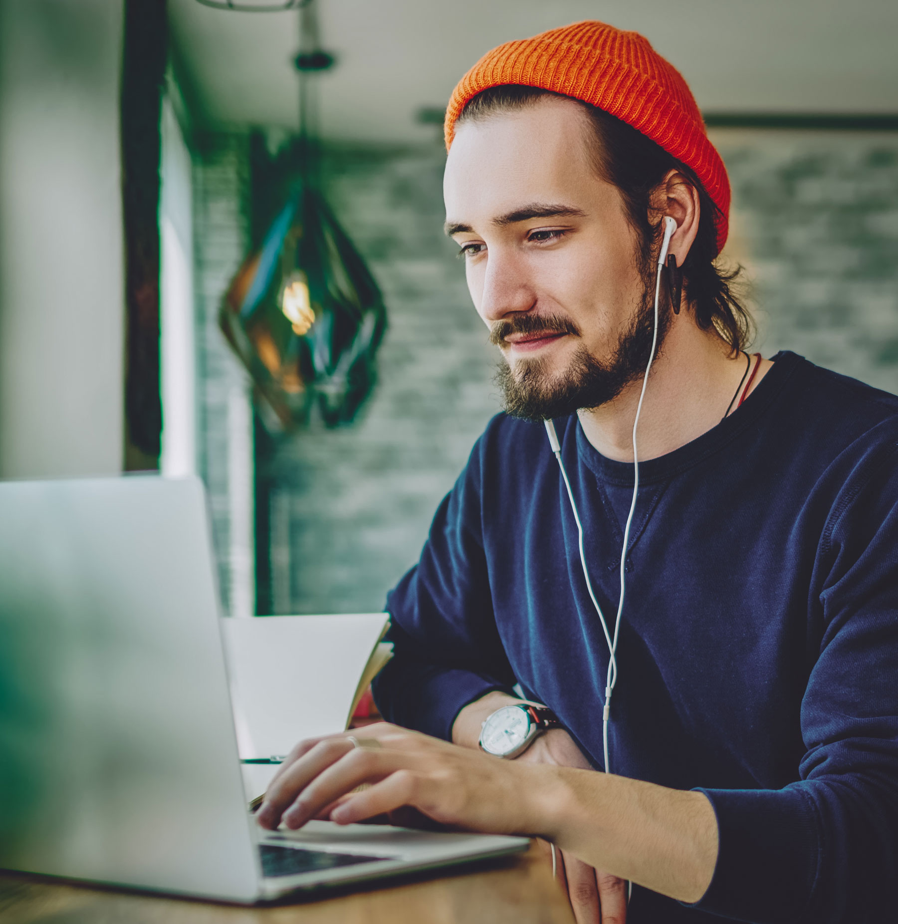 Young man in coffee shop looking a laptop with earbuds and a beanie