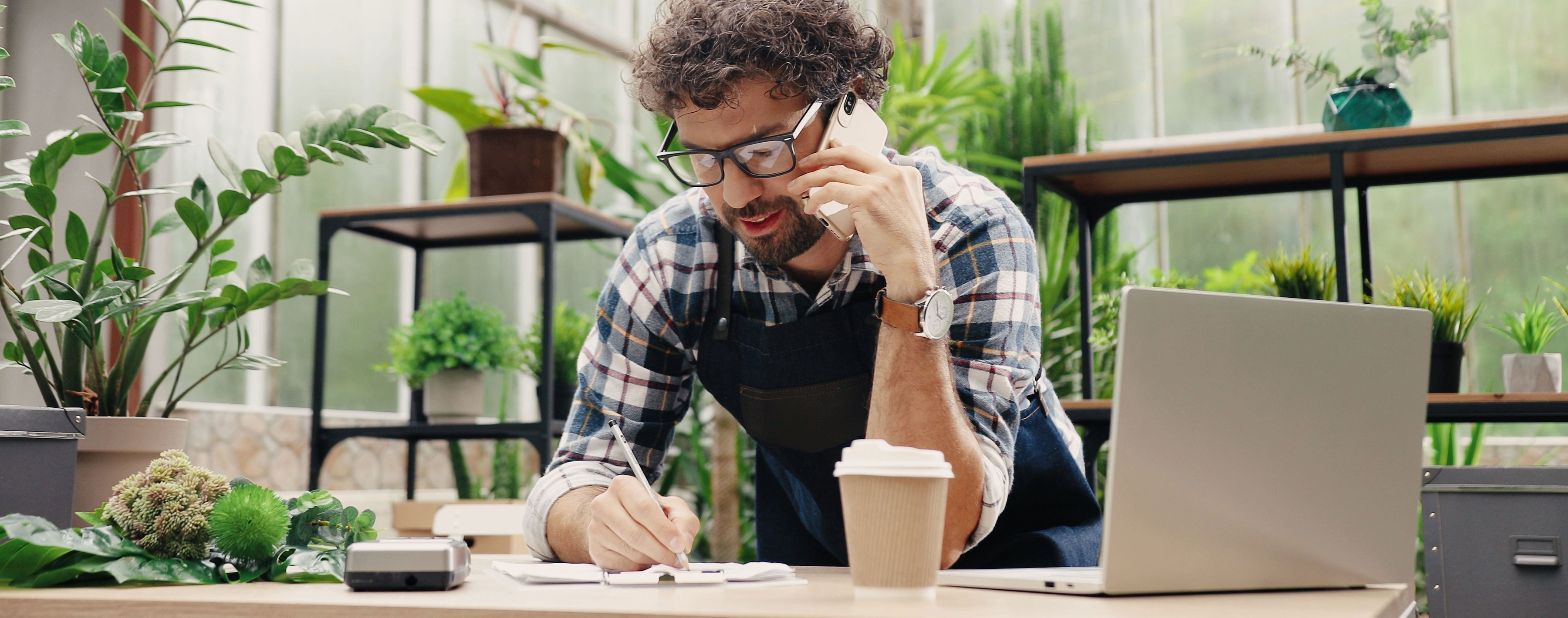 Happy Caucasian businessman talking on cellphone while standing in apron in small floral center and writing down order details. Joyful male florist calling on smartphone at work.